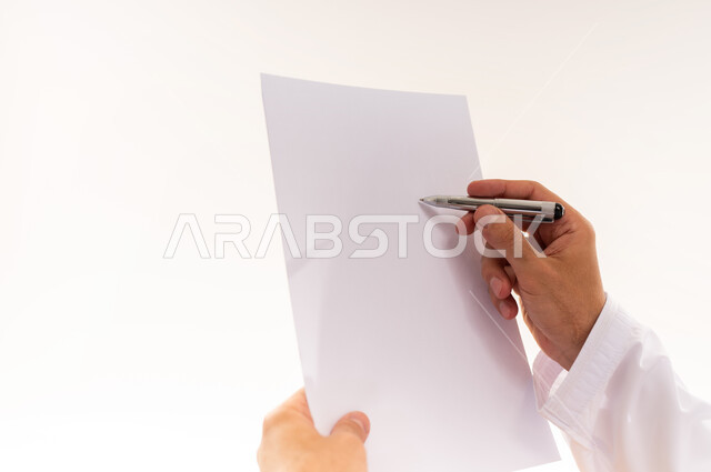 القيام بأداء المهام اليومية المكتبيه, بورتريه مقرب لأيدي رجل عربي خليجي عماني يحمل بيده قلم وورقة يشير الى شيء محدد, مهنة ووظيفة شبابية, التدقيق على التقرير الشهري, خلفية بيضاء