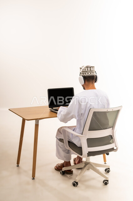 مهنة ووظيفة مكتبية، مفهوم الإصغاء والإستماع، عرض اللابتوب المحمول بشاشة سوداء فارغة، دمج التقنية والتكنولوجيا المتطورة بالعمل، بورتريه من الخلف لرجل عربي خليجي عماني يرتدي الدشداشة والكمه يعمل على جهاز الحاسوب بجانب سماعة الهيدفون، خلفية بيضاء