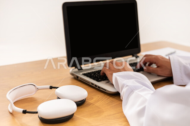 مفهوم الإصغاء والإستماع، دمج التقنية والتكنولوجيا المتطورة بالعمل، مهنة ووظيفة مكتبية، عرض اللابتوب المحمول بشاشة سوداء فارغة، بورتريه مقرب لأيدي رجل عربي خليجي عماني يعمل على جهاز الحاسوب بجانب سماعة الهيدفون، خلفية بيضاء