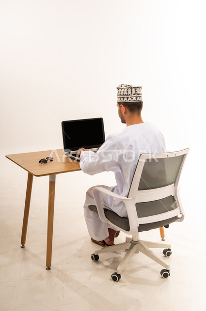 مهنة ووظيفة مكتبية، عرض اللابتوب المحمول بشاشة سوداء فارغة، دمج التقنية والتكنولوجيا المتطورة بالعمل، بورتريه من الخلف لرجل عربي خليجي عماني يرتدي الدشداشة والكمه يجلس على كرسي مريح يعمل على جهاز الحاسوب بجانب النظارة الشمسية، خلفية بيضاء