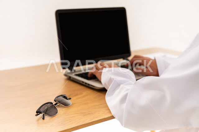 دمج التقنية والتكنولوجيا المتطورة بالعمل، مهنة ووظيفة مكتبية، عرض اللابتوب المحمول بشاشة سوداء فارغة، بورتريه مقرب لأيدي رجل عربي خليجي عماني يعمل على جهاز الحاسوب بجانب النظارة الشمسية، خلفية بيضاء