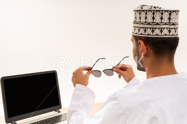 Laptop display with blank black screen, technology and advanced technology integration in work, portrait from back of an Arab Gulf Omani man wearing a dishdasha and a kimono holding sunglasses in his hand, office profession and job, white background