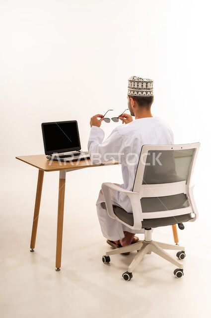 مهنة ووظيفة مكتبية، عرض اللابتوب المحمول بشاشة سوداء فارغة، دمج التقنية والتكنولوجيا المتطورة بالعمل، بورتريه من الخلف لرجل عربي خليجي عماني يرتدي الدشداشة والكمه يجلس على كرسي مريح يمسك بيده النضارة الشمسية، خلفية بيضاء