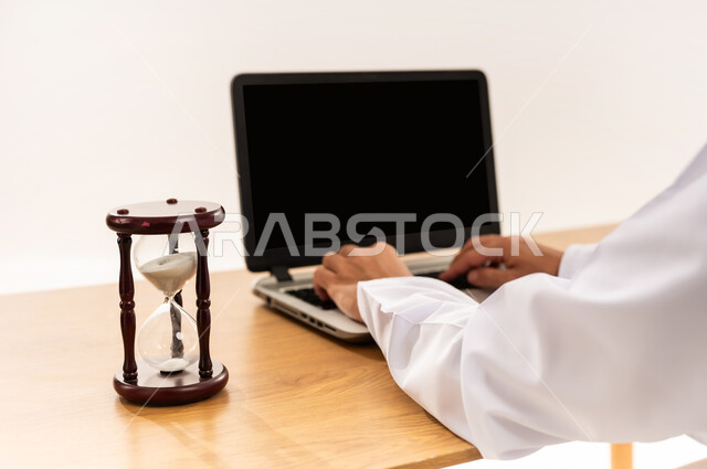 دمج التقنية والتكنولوجيا المتطورة بالعمل، بورتريه مقرب لأيدي رجل عربي خليجي عماني يعمل على جهاز اللابتوب المحمول بجانب الساعة الرملية, انجاز المهام بدقة وكفاءة، ادارة الجدول الزمني، عرض اللابتوب المحمول بشاشة سوداء فارغة، خلفية بيضاء