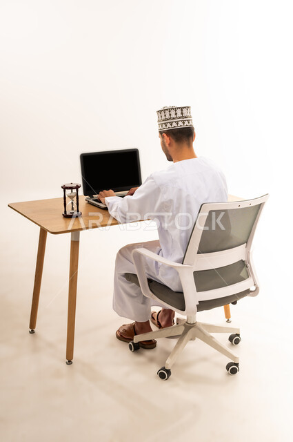 انجاز المهام بدقة وكفاءة، دمج التقنية والتكنولوجيا المتطورة بالعمل، بورتريه من الخلف لرجل عربي خليجي عماني يرتدي الدشداشة والكمه يعمل على جهاز اللابتوب المحمول بجانب الساعة الرملية, ادارة الجدول الزمني، عرض اللابتوب المحمول بشاشة سوداء فارغة، خلفية بيضاء