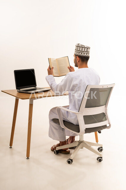 مراجعة الدروس اليومية، مهنة و عمل مكتبي إداري, دمج التقنية الحديثة و المتطورة مع العمل, بورتريه من الخلف لرجل عربي خليجي عماني يرتدي الدشداشة والكمه يحمل بيده كتاب يقوم بالتحضير للاختبار النهائي, عرض اللابتوب المحمول بشاشة سوداء فارغة، خلفية بيضاء