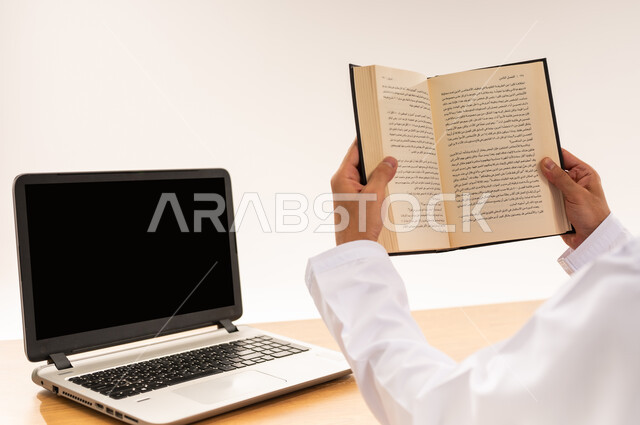 التحضير للاختبار النهائي، مهنة و عمل مكتبي إداري, دمج التقنية الحديثة و المتطورة مع العمل, بورتريه مقرب لأيدي رجل عربي خليجي عماني يرتدي الدشداشة والكمه يحمل بيده كتاب يقوم بمراجعة الدروس اليومية, عرض اللابتوب المحمول بشاشة سوداء فارغة، خلفية بيضاء