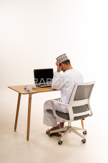 الشعور بالتوتر والصداع، مهنة و عمل مكتبي إداري, دمج التقنية الحديثة و المتطورة مع العمل, بورتريه من الخلف لأيدي رجل عربي خليجي عماني يرتدي الدشداشة والكمه يضع يده على رأسه بإيماءات التعب والإرهاق, عرض اللابتوب المحمول بشاشة سوداء فارغة، خلفية بيضاء