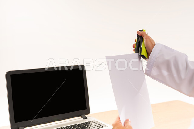 مهنة و عمل مكتبي إداري, دمج التقنية الحديثة و المتطورة مع العمل, إعداد التقرير الشهري, بورتريه مقرب من الخلف لرجل عربي خليجي عماني يرتدي الكمه و الدشداشة يجلس على كرسي يقوم بخرز الورق، عرض اللابتوب المحمول بشاشة سوداء فارغة, خلفية بيضاء