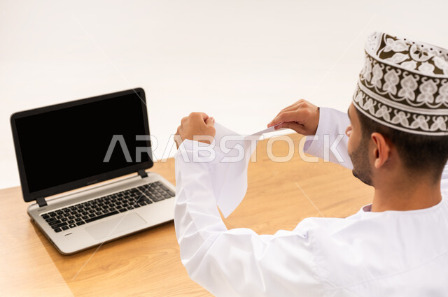 اتلاف بعض المعلومات القديمة الغير مهمة, عدم الموافقة على خطة عمل فاشلة, بورتريه مقرب من الخلف لرجل عربي خليجي عماني يرتدي الكمه و الدشداشة يجلس على الكرسي أمام طاولة خشبية يقوم بتمزيق الاوراق، صفقة غير ناجحة، خلفية بيضاء