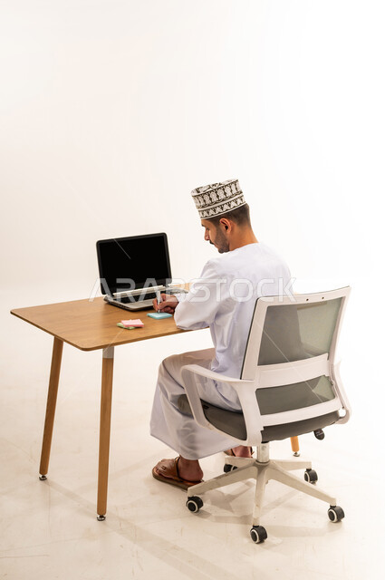 وظيفة ومهنة عمانية شبابية, استخدام جهاز تقني حديث متطور, بورتريه من الخلف لرجل عربي خليجي عماني يرتدي الكمه و الدشداشة يجلس على الكرسي أمام طاولة خشبية يقوم بتدوين الأفكار وتسجيل المعلومات على الأوراق الملونة, خلفية بيضاء