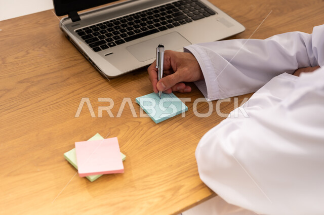 استخدام جهاز تقني حديث متطور, مهنة ووظيفة عمانية شبابية, بورتريه مقرب من الخلف لرجل عربي خليجي عماني يرتدي الزي التقليدي يجلس على طاولة خشبية يقوم بتدوين الأفكار وتسجيل المعلومات على الأوراق الملونة, خلفية بيضاء