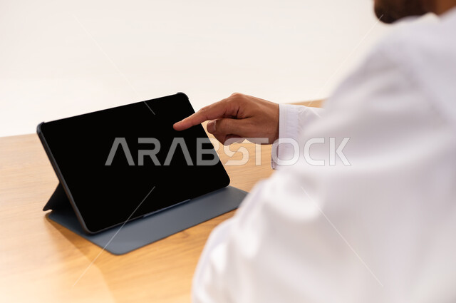 التواصل و الدردشة مع الاقارب, القيام بمقابلات وإجتماعات العمل أونلاين، استخدام جهاز تقني حديث لتصفح الصور والفيديوهات بسهولة، بورتريه مقرب من الخلف لرجل عربي خليجي عماني يرتدي الزي التقليدي يضغط باصبع السبابة على جهاز التابلت بشاشه سوداء فارغة, خلفية بيضاء