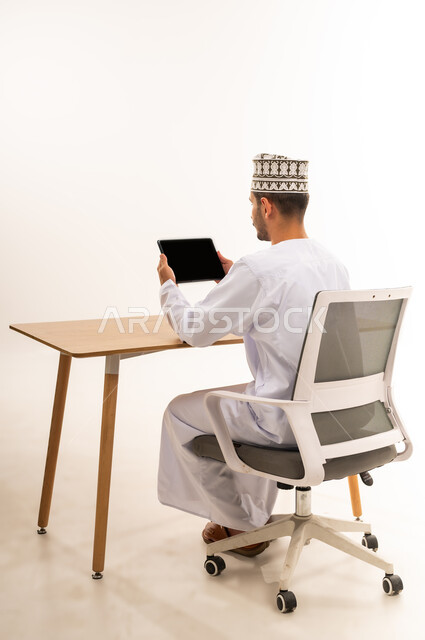 التطور التقني في مختلف المجالات, استخدام التكنولوجيا الحديثة في العمل عن بعد، عقد الاجتماعات عن طريق الانترنت، إنجاز الأعمال بسرعة وسهولة، بورتريه من الخلف لرجل عربي خليجي عماني يرتدي الكمه و الدشداشة يحمل بيده جهاز التابليت بشاشة سوداء فارغة, خلفيه بيضاء