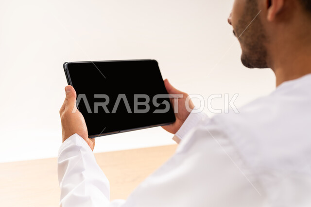 استخدام التكنولوجيا الحديثة في العمل عن بعد، عقد الاجتماعات عن طريق الانترنت، إنجاز الأعمال بسرعة وسهولة، التطور التقني في مختلف المجالات، بورتريه مقرب من الخلف لرجل عربي خليجي عماني يرتدي الزي التقليدي يحمل بيده جهاز التابليت بشاشة سوداء فارغة, خلفيه بيضاء