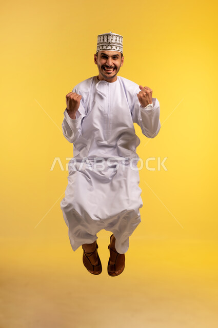 ايماءات الفوز و الكسب, الرضا و الشعور بالاعجاب، تحقيق الاهداف و إنجاز المهامات ,بورتريه لرجل عربي خليجي عماني يرتدي الكمه و الدشداشة يقفز عاليا يرفع قبضه يده بتعابير الفرح بالنصر, علامة السعادة والقوه, خلفية صفراء