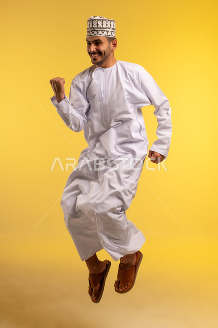 الرضا و الشعور بالاعجاب، تحقيق الاهداف و إنجاز المهامات ,بورتريه لرجل عربي خليجي عماني يرتدي الكمه و الدشداشة يقفز عاليا يرفع قبضه يده بتعابير الفرح بالنصر, ايماءات الفوز و الكسب, علامة السعادة والقوه, خلفية صفراء