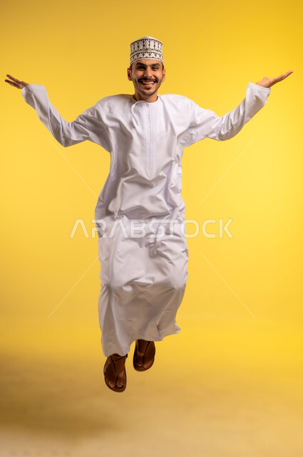 عدم التأكد والقدرة على اتخاذ القرار، التردد بشأن أمر معين، بورتريه لرجل عربي خليجي عماني مبتسم يرتدي الكمه و الدشداشة يقفز عاليا يرفع كفيه نحو الأعلى بتعابير الحيرة، النظر إلى الكاميرا بإيماءات الاستغراب، صورة الجسم بالطول الكامل، خلفية صفراء