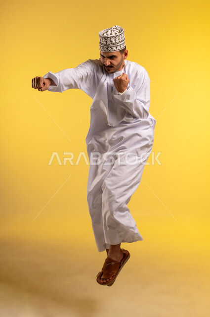 ممارسة رياضة الدفاع عن النفس، تعزيز صحة العظام والمفاصل، إتقان فنون الملاكمة، زيادة قدرة العضلات على التحمل، بورتريه لرجل عربي خليجي عماني يرتدي الكمه و الدشداشة يقفز عاليا يرفع قبضات يديه بإيماءات القوة، صورة الجسم بالطول الكامل, خلفية صفراء