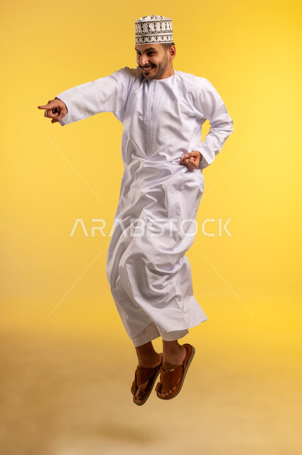 الاهتمام بالمظهر الخارجي, النظر الى شيء ما بايماءات الفرح والسرور, بورتريه لشاب عربي خليجي عماني مبتسم يرتدي الكمه و الدشداشة يقفز عاليا يشير بأصبع السبابة إلى شيء ما, صورة الجسم بالطول الكامل، خلفية صفراء