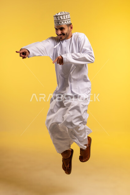 الاهتمام بالمظهر الخارجي، الاشارة بأصابع السبابة إلى شيء ما، بورتريه لشاب عربي خليجي عماني مبتسم يرتدي الكمه و الدشداشة يقفز عاليا وينظر الى شيء ما بايماءات الفرح والسرور, صورة الجسم بالطول الكامل، خلفية صفراء