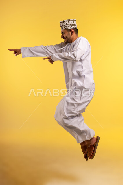 Pointing with index fingers at something, concern for appearance, side portrait of a young Arab Gulf Omani man wearing a kimono and a dishdasha jumping high and looking at something with gestures of joy and happiness, full body image, yellow background