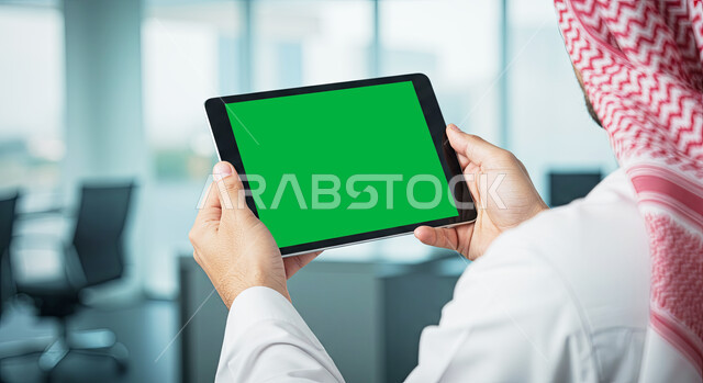 عرض تابلت بشاشة خضراء فارغة، تصفح مواقع التواصل الاجتماعي عن طريق الانترنت، استخدام الأجهزة الحديثة في انجاز الأعمال، استعمال التكنولوجيا في العمل عن بعد، صورة مقربة من الخلف لرجل عربي خليجي سعودي يرتدي الثوب التقليدي والشماغ يحمل بيده جهاز لوحي في المكتب، كروما