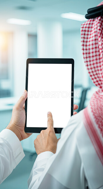 متابعة مواقع التواصل الاجتماعي عن طريق الانترنت، عرض تابلت بشاشة بيضاء فارغة، استخدام الأجهزة الحديثة في انجاز الأعمال، استعمال التكنولوجيا في العمل عن بعد، صورة مقربة من الخلف لرجل عربي خليجي سعودي يرتدي الثوب التقليدي والشماغ يحمل بيده جهاز لوحي في المكتب