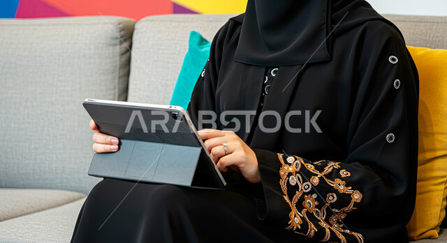 تصفح مواقع التواصل الاجتماعي، مفهوم إنجاز المشاريع ومهام العمل عن بعد، استخدام الأجهزة التقنية الحديثة، التواصل مع الأهل والأصدقاء عبر الايباد، صورة مقربة لإمرأة عربية خليجية سعودية محجبة ترتدي العباءة السوداء تجلس على اريكة مريحة تقوم باستخدام الجهاز اللوحي