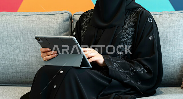 مفهوم إنجاز المشاريع ومهام العمل عن بعد، استخدام الأجهزة التقنية الحديثة، تصفح مواقع التواصل الاجتماعي، التواصل مع الأهل والأصدقاء عبر الايباد، صورة مقربة لإمرأة عربية خليجية سعودية محجبة ترتدي العباءة السوداء تجلس على اريكة مريحة تقوم باستخدام الجهاز اللوحي
