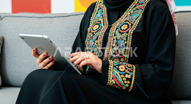 Using modern technical devices, the concept of completing projects and tasks, working remotely, browsing social networking sites, communicating with family and friends via iPad, a close-up image of a veiled Saudi Arabian Gulf woman wearing a black abaya sitting on a comfortable sofa using a tablet