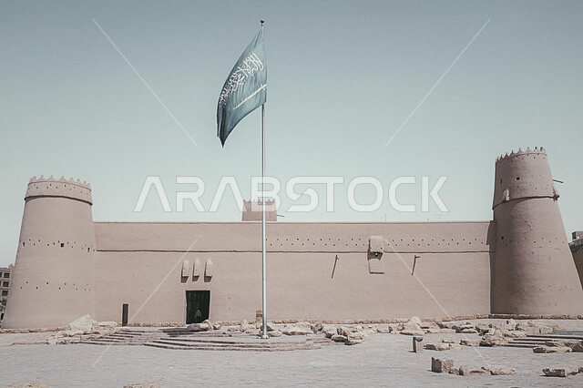 سارية العلم الوطني السعودي يرفرف بالسماء، قصر المصمك التاريخي في مدينة الرياض في المملكة العربية السعودية، الهندسة المعمارية للمباني الطينية القديمة من التراث السعودي، مكان ومعلم أثري سياحي في السعودية