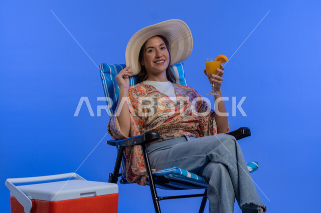 تناول المشروبات الباردة المنعشة، شابة سعودية تجلس على الكرسي للاسترخاء، بورتريه لإمرأة عربيه خليجية اماراتية مبتسمه ترتدي الزي الكاجوال و قبعة القش العصرية بايماءات السرور والاستمتاع، قضاء اجازة الصيف بالاماكن السياحية, خلفية زرقاء