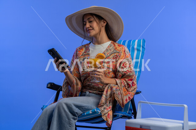 Having a nice vacation, a Saudi woman taking selfies with her mobile phone, drinking refreshing natural juices, close-up portrait of a smiling young Arab Emirati woman in casual wear and a straw hat using a mobile phone, enjoying a summer vacation, blue background