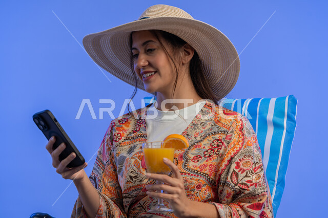 قضاء أيام ممتعة في العطلة, إمرأة سعودية تقوم بالتقاط صور سيلفي تذكارية عبر الجوال, شرب العصائر الطبيعيه المنعشة, بورتريه مقرب لشابة عربية خليجية اماراتية مبتسمة ترتدي الزي الكاجوال والقبعة القش تستخدم الهاتف المحمول, الاستمتاع بالإجازة الصيفية, خلفية زرقاء