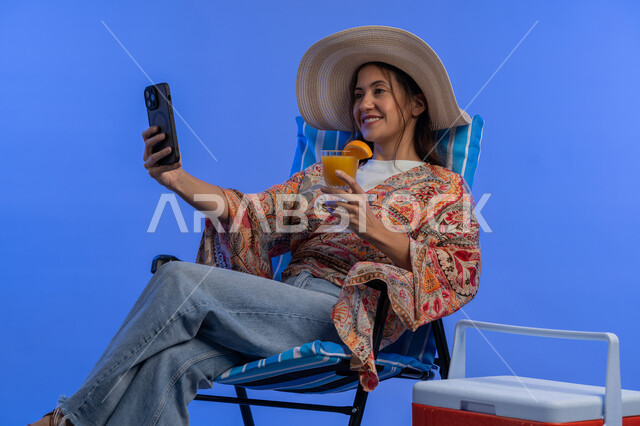 Having a nice vacation, a Saudi woman taking selfies with her mobile phone, drinking refreshing natural juices, close-up portrait of a smiling young Arab Emirati woman in casual wear and a straw hat using a mobile phone, enjoying a summer vacation, blue background