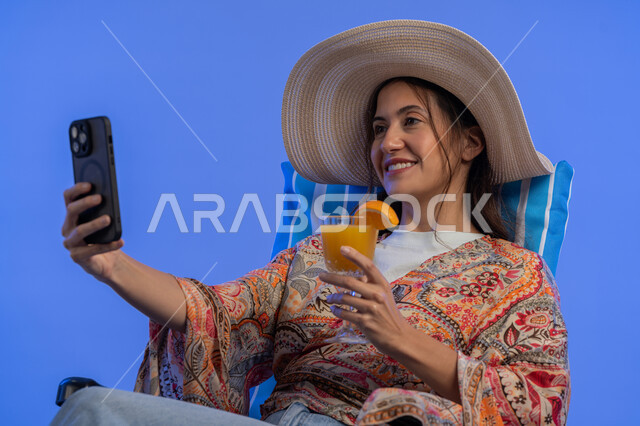 قضاء أيام ممتعة في العطلة, إمرأة سعودية تقوم بالتقاط صور سيلفي تذكارية عبر الجوال, شرب العصائر الطبيعيه المنعشة, بورتريه مقرب لشابة عربية خليجية اماراتية مبتسمة ترتدي الزي الكاجوال والقبعة القش تستخدم الهاتف المحمول, الاستمتاع بالإجازة الصيفية, خلفية زرقاء