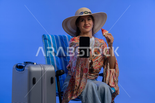 Eating refreshing summer fruits, using mobile travel and tourism apps, enjoying summer vacation, close-up portrait of young Arab Gulf Emirati woman showing blank black mobile phone screen, Saudi woman sitting on chair to relax, blue background