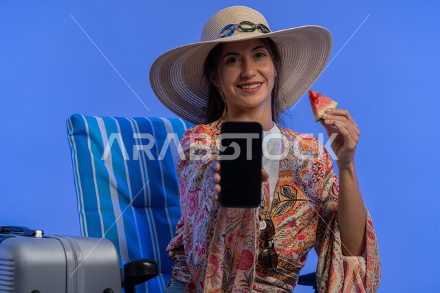 Eating refreshing summer fruits, using mobile travel and tourism apps, enjoying summer vacation, close-up portrait of young Arab Gulf Emirati woman showing blank black mobile phone screen, Saudi woman sitting on chair to relax, blue background