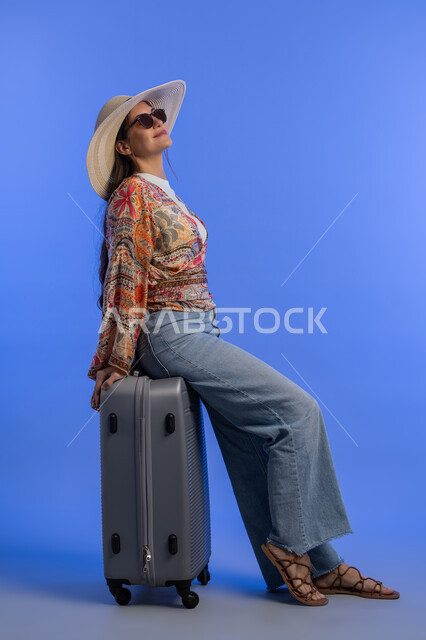 مفهوم اللطافة والأنوثة والنعومة، التسلية والترفيه في الخارج، إمرأة سعودية تقوم بالاستعداد للسفر الممتع, بورتريه لشابة عربية خليجية اماراتية مبتسمة ترتدي الزي الكاجوال مع قبعة القش تجلس على حقيبة الأمتعة بإيماءات السرور والمتعة والحماس, خلفية زرقاء