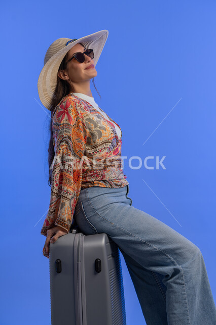 مفهوم اللطافة والأنوثة والنعومة، التسلية والترفيه في الخارج، إمرأة سعودية تقوم بالاستعداد للسفر الممتع, بورتريه لشابة عربية خليجية اماراتية مبتسمة ترتدي الزي الكاجوال مع قبعة القش تجلس على حقيبة الأمتعة بإيماءات السرور والمتعة والحماس, خلفية زرقاء