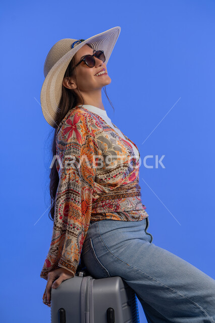 مفهوم اللطافة والأنوثة والنعومة، التسلية والترفيه في الخارج، إمرأة سعودية تقوم بالاستعداد للسفر الممتع, بورتريه لشابة عربية خليجية اماراتية مبتسمة ترتدي الزي الكاجوال مع قبعة القش تجلس على حقيبة الأمتعة بإيماءات السرور والمتعة والحماس, خلفية زرقاء