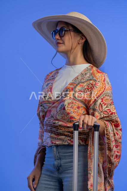 التسلية والترفيه في الخارج، مفهوم اللطافة والأنوثة والنعومة، إمرأة سعودية تقوم بالاستعداد للسفر الممتع, بورتريه لشابة عربية خليجية اماراتية مبتسمة ترتدي الزي الكاجوال والنظارات الشمسية مع قبعة القش تجر حقيبة الأمتعة بإيماءات السرور والمتعة والحماس, خلفية زرقاء
