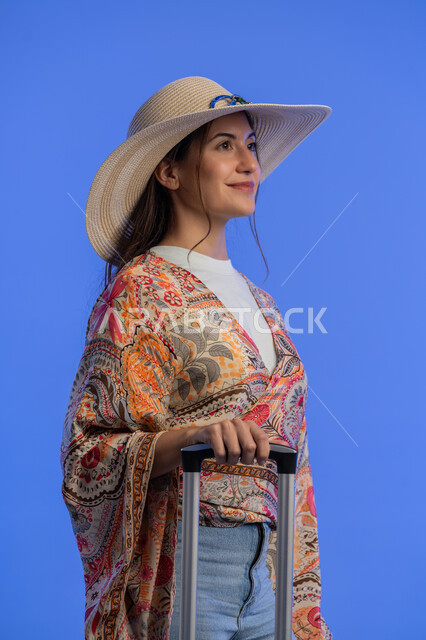 Entertainment and leisure abroad, the concept of gentleness, femininity and softness, a Saudi woman preparing for an enjoyable trip, a portrait of a smiling young Arab Gulf Emirati woman wearing casual clothes with a straw hat pulling her luggage with gestures of pleasure, enjoyment and enthusiasm, blue background