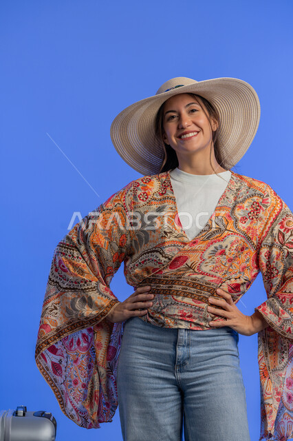 Gestures of pleasure, enjoyment and enthusiasm, entertainment and recreation abroad, concept of gentleness, femininity and softness, Saudi woman preparing for an enjoyable trip, portrait of a smiling young Arab Gulf Emirati woman wearing casual clothes with a straw hat standing next to a luggage bag, blue background