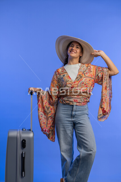 Gestures of pleasure, enjoyment and enthusiasm, entertainment and recreation abroad, concept of gentleness, femininity and softness, Saudi woman preparing for an enjoyable trip, portrait of a smiling young Arab Gulf Emirati woman wearing casual clothes with a straw hat standing next to a luggage bag, blue background