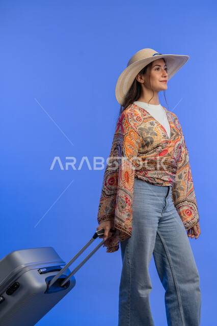 Gestures of pleasure, enjoyment and enthusiasm, entertainment and recreation abroad, the concept of gentleness, femininity and softness, a Saudi woman preparing for an enjoyable trip, a portrait of a smiling young Arab Gulf Emirati woman wearing casual clothes with a straw hat pulling a luggage bag, blue background