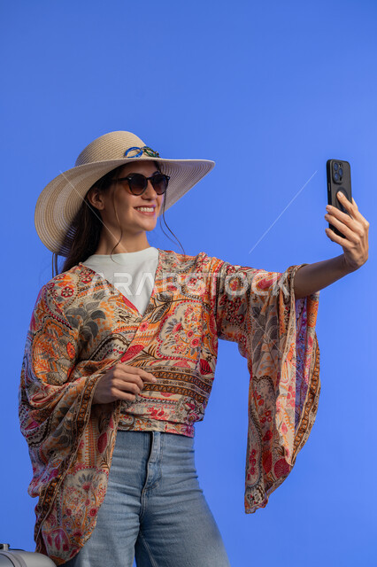 توثيق اللحظات الممتعه باستخدام الجوال, الاهتمام بالأزياء والموضة، بورتريه لشابة عربية خليجية اماراتية مبتسمة ترتدي الزي الكاجوال والنظارة الشمسية مع قبعة القش تقوم بالتقاط صور سيلفي تذكارية عبر الهاتف المحمول، إمرأة سعودية تستخدم التقنية والتكنولوجيا, خلفية زرقاء