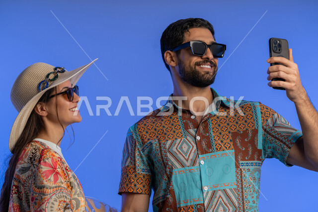 استخدام الهاتف المحمول لالتقاط صور السيلفي التذكارية, قضاء وقت ممتع على الشاطئ, بورتريه مقرب لزوجيين عربين خليجيين اماراتيين مبتسمين يرتديان الزي الكاجوال يقومان بتوثيق اللحظات السعيدة, عائلة سعودية تقف بإيماءات السرور والسعادة,  خلفية زرقاء