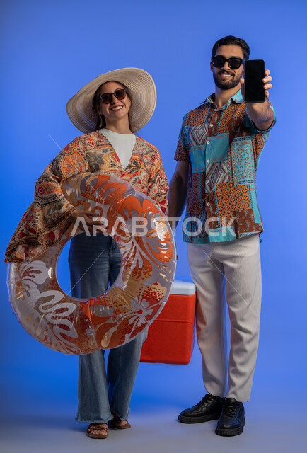 عرض شاشة سوداء فارغة على الهاتف المحمول, الاستمتاع والترفيه على الشواطئ, بورتريه لزوجيين عربين خليجيين اماراتيين مبتسمين يرتديان الزي الكاجوال والنظارة الشمسية يقضيان اوقات ممتعة في العطلة, شابة سعودية تحمل بيدها عوامة السباحة، ​الجسم بالطول الكامل, خلفية زرقاء